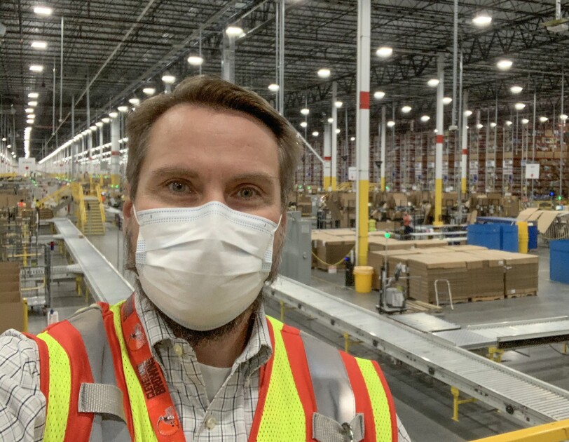 A man wearing a protective mask and a safety vest stands in front of a large warehouse space.