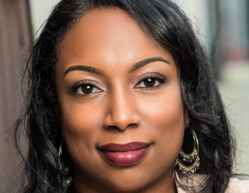 A woman with dark skin and black hair looks at the camera with a slight smile on her face