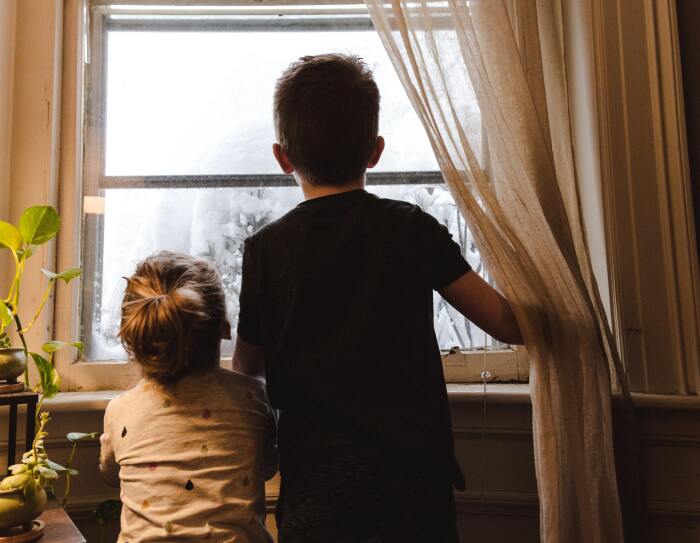 Doa heramos: una hermana de unos 3 años y un hermano de unos 7 años. Los dos están de espaldas y mirando por una ventana. El niño va con una camiseta negra de manga corta. La niña, con una camiseta blanca y con lágrimas de colores. Fuera de la ventana se ve todo blanco y parece que hay un árbol con nieve. El niño con la mano derecha aguanta unas cortinas blancas. El marco d e la ventana es blanco y de madera. En laparte izquierda de la foto hay un mueble con 4 plantes y se nota que entra la luz de una lámpara.