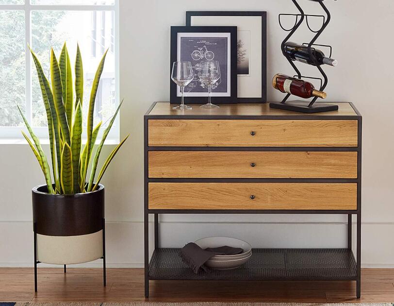 A raised pot on three feet holds a tall green plant. To the right of the plant is a three drawer sideboard with a wine rack, two wine glasses and framed art on top of it. In front the sideboard and plant is a woven rug.
