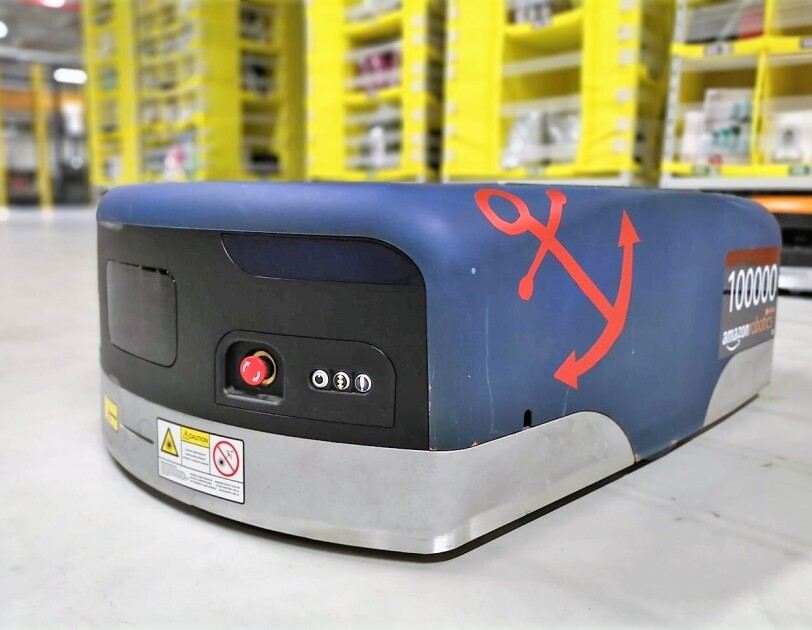 Auf dem Boden einer Lagerhalle sieht man einen blauen Amazon Transportroboter. Auf der rechten Seite wurde ein roter Anker aufgeklebt und ebenfalls rechts steht die kennzeichnung "100000 amazon robotics".