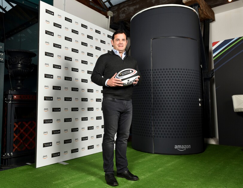 Will Carling standing holding a rugby ball at the Six Nations promoting AWS. He is standing in front of a large Amazon Alexa Echo model.