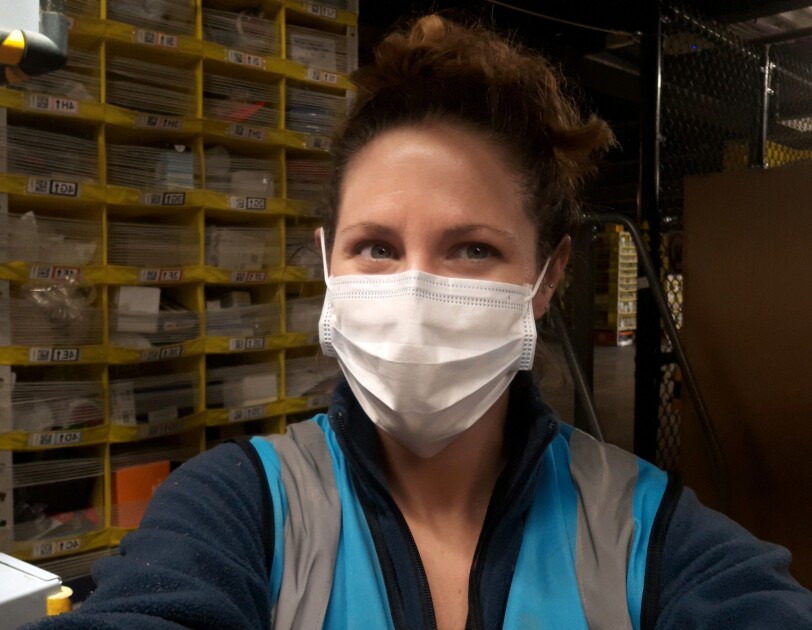 A woman in a safety mask stands with shelving behind her.