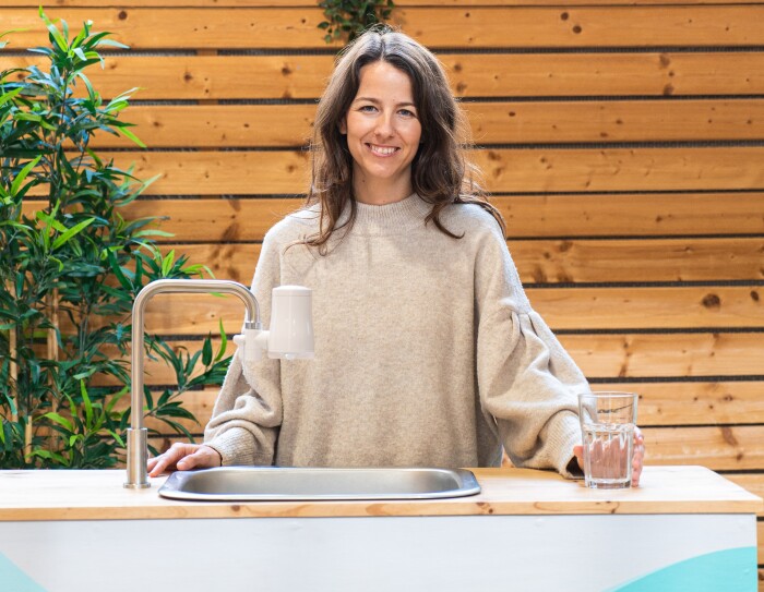La start-up TAPP Water fue fundada por Magnus Jern y Alexander Schwarz. En la foto aparece la CEO Rocío Alcocer en una mesa con un gripo que tiene el filtro de Tapp water . Rocío va un jersey también marrón muy claro. Tiene el pelo largo castaño y las manos apoyadas sobre la mesa, sujerta una vaso de cristal con agua. De fondo una pared de madera.