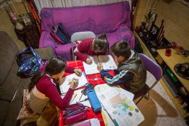 Tres niños, dos niñas y un niños, trabajando en una mesa situada en el salón de una casa. Dos de ellos están sentados enuna silla y la tercera está sentada en el sofà. Dos de ellos trabajan sobre el mismo libro. Hay dos sofás y en la mesa hay libros, lápices y rotuladores. Los tres van muy abrigados y tienen las mochilas tiradas por el sofá.