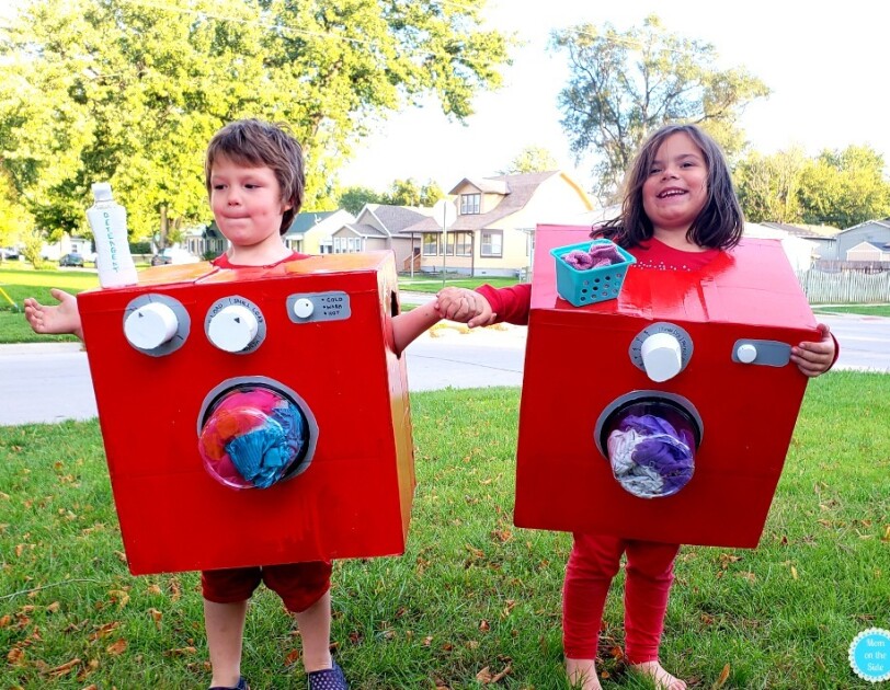Two young children stand side-by-side, each wearing a red box. Both boxes have a clear "window" on the front that shows clothing, which look as though they're tumbling in the laundry. The child on the left has a bottle that says "detergent" on the top of the box, and two knobs. The child on the right has a small basket that looks like a laundry basket with socks, and one knob.