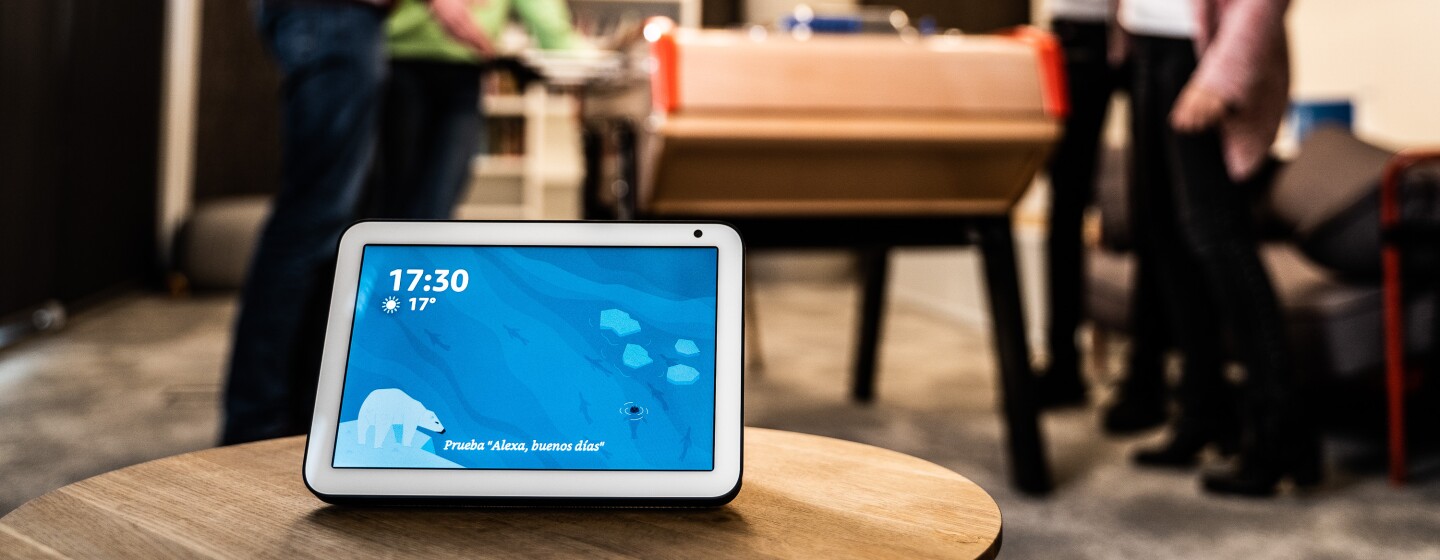 El dispositivo Echo Show en primer plano con la pantalla de color azul la hora y la temperatura. EL dispositivo se encuentra encima de una mesa. De fondo se ven cuatro personas jugando al futbolín. Dos chicos y dos chicas. Las parejas son mixtas y una de ellas se está celebrando que ha marcado un gol.