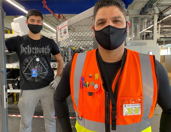 Daniel Fonseca y Diego Fonseca en el centro logístico de Amazon en El Prat de Llobregat (Barcelona). De fondo unas cajas de color negro y el techo con conductos de color azul. En primer plano Daniel con una camiseta térmica estrecha negra de manga larga y un chaleco naranja. Mascarilla de color negro, pelo negro corto y mirando a cámara. En segundo plano está Diego, con un pantalón de deporte una camiseta negra de manga corta. Lleva guantes y la mano izauierda la apoya en la cintura y la derecha en una puerta metálica. Mascarilla negra.
