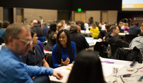 Diverse woman seated at a table in a conference room, is engaging in conversation with other conference attendees.