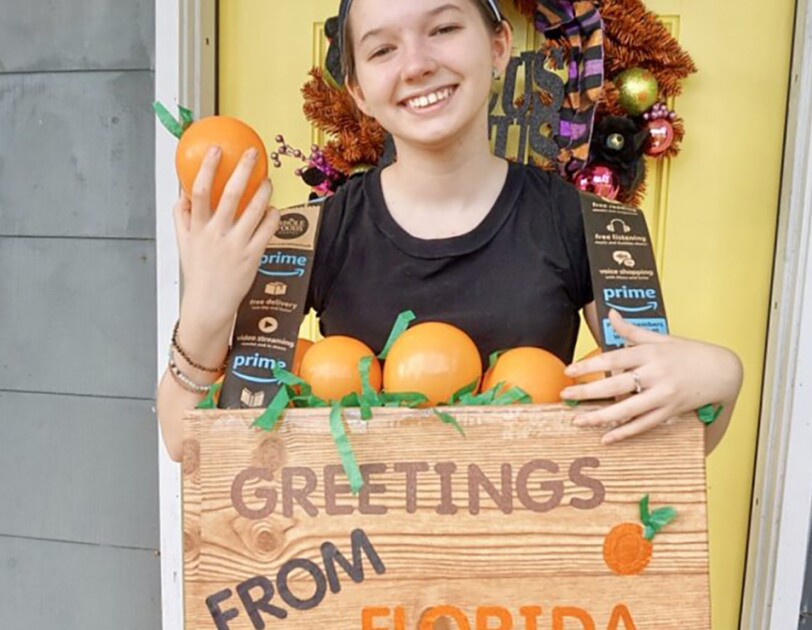 A young woman wears a Halloween costume that is made of a box around her torso, in wood grain, that says "greetings from Florida" on the front. The top of the box is open and shows fake grass and pretend oranges, as though it was a fruit box.