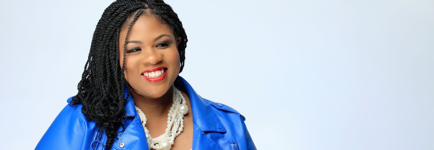 Amazonian con un fondo blanco y un primer plano hasta la cintura, Mujer con trenzas negras, maquillaje en los labios rojos y los ojos negros. Lleva un collar de perlas de diferentes tamaños, una chaqueta como de piel de color azul eléctrico y un jersei blanco roto estrecho. Tiene también las uñas de color rojo. 