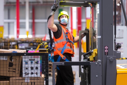 Dipendente di un magazzino Amazon. Indossa un casco, una mascherina chirurgica e un giubbino di sicurezza, si trova al centro di un macchinario. Alle sue spalle un cesto con dei pacchi Amazon.
