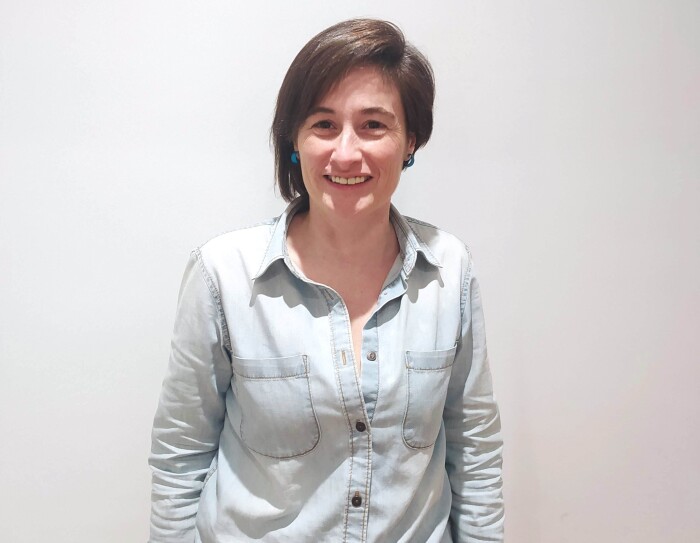Marta Casasayas. Detrás un fondo blanco de pared. Marta aparece en un plano hasta los muslos. Lleva una camisa tejana azul clara de manga larga y unos jeans azul oscuro. Tiene el pelo corto y la parte izquierda más larga que la derecha. Está sonriendo y lleva unos pendientes que osn unos aros de color azul.