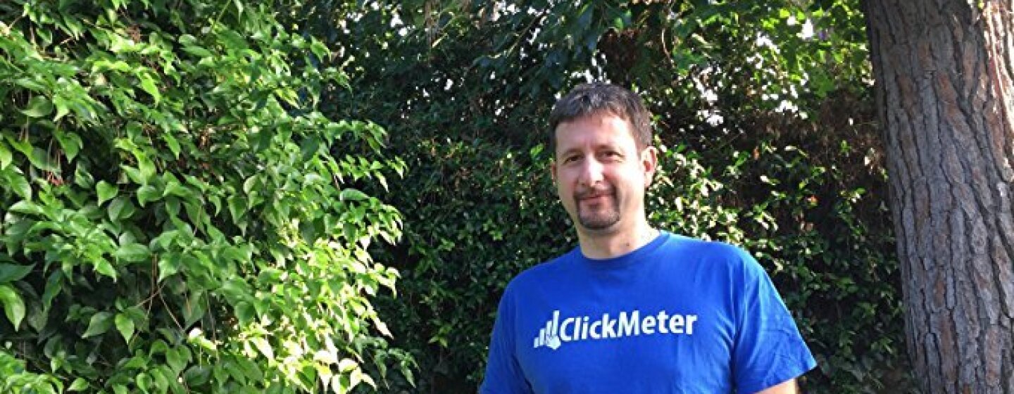 Ritratto a mezzo busto di Davide De Guz, sviluppatore di ClickMeter. L'uomo si trova in un giardino e indossa una t-shirt blu con il logo della sua azienda.