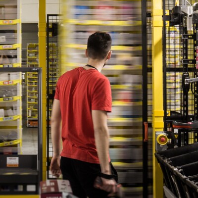 Un dipendente Amazon ripreso di spalle al centro dell'immagine osserva gli scaffali mobili del magazzino.