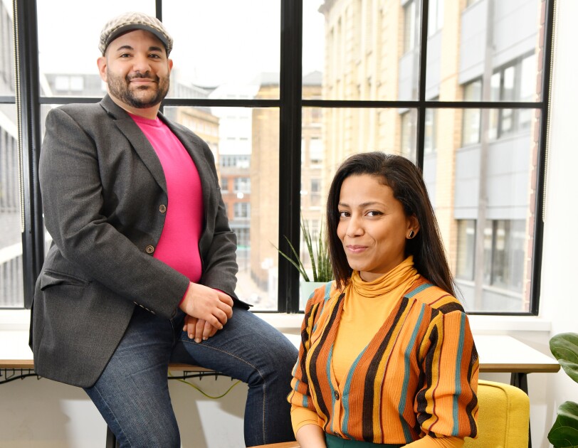 A portrait of Naveed and Samiya Parvez. Naveed is leaning against a window in their Shoreditch office, and Samiya is sitting on a rocking chair.