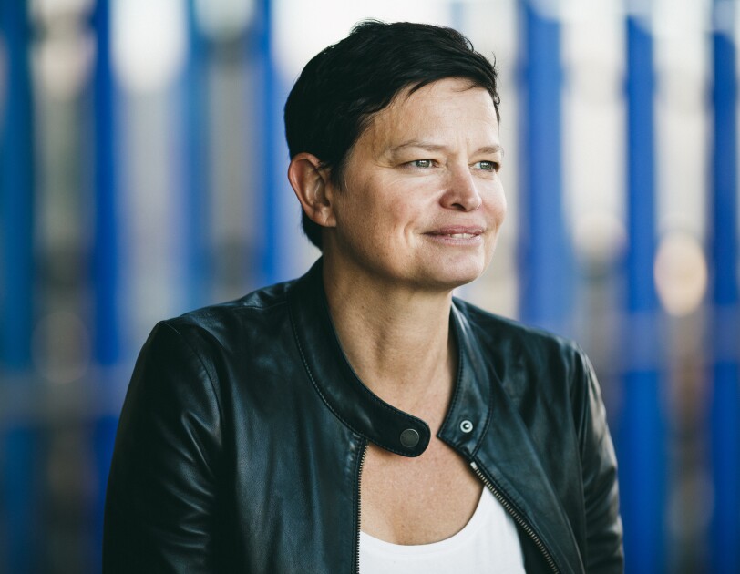 A dark-haired woman, Amazon's disaster relief head Bettina Stix, is dressed in a black leather jacket looks to the right and smiling. There is a blue background.
