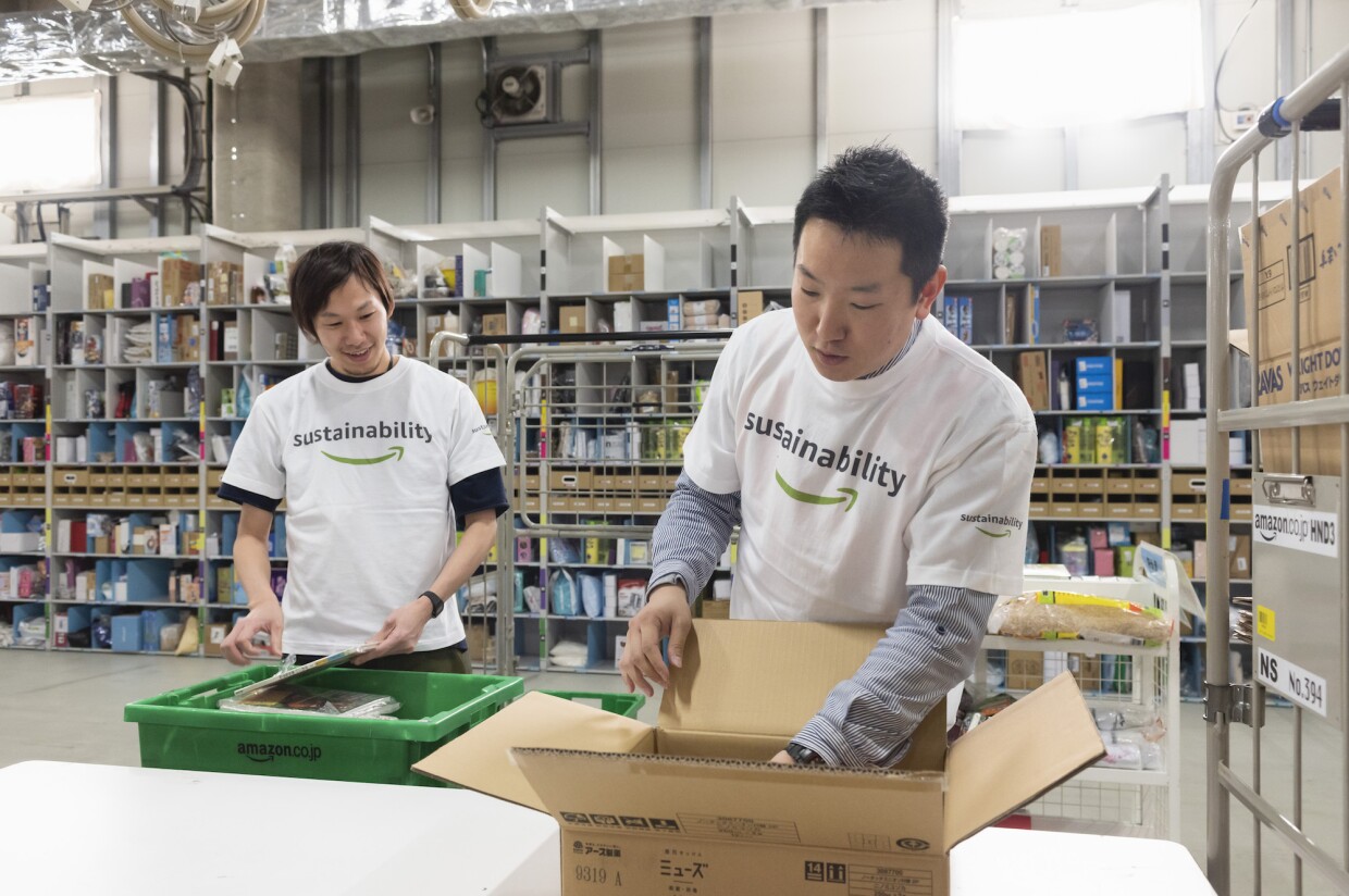 Two Amazon employees wearing 'sustainability' t-shirts work to pack a box together.