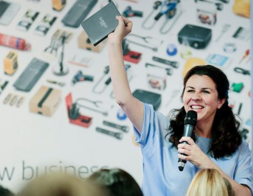 Woman with brown hair holding a mic and talking to an audience