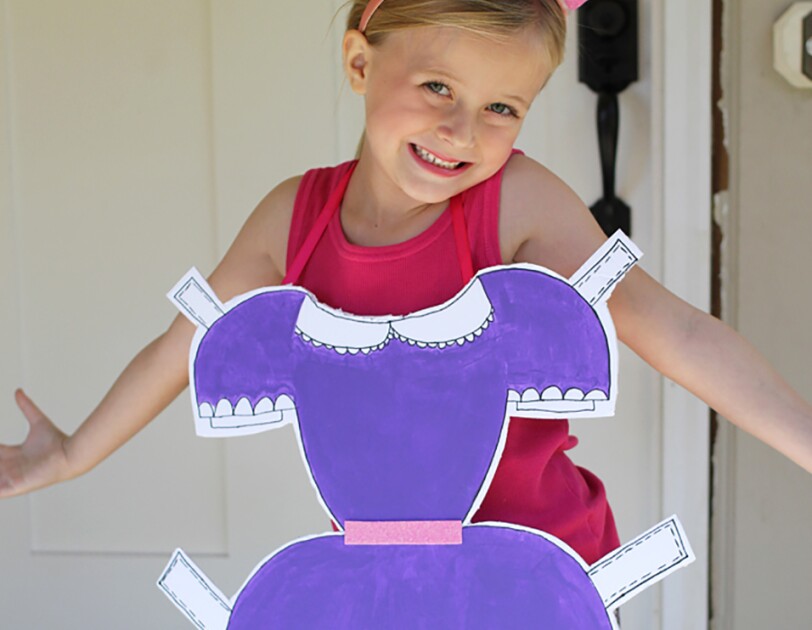 A little girl in a "paper doll" costume. She wears a tank top and leggings, and wears a dress with tabs (like a paper doll dress) on her front.