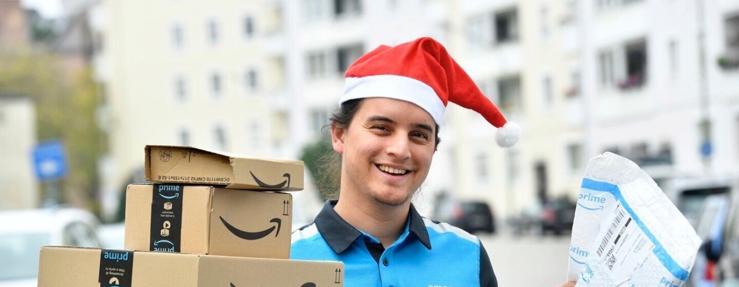 Ein junger Mann in Amazon Zustellkleidung und roter Zipfelmütze hält eine Reihe von Paketen in der Hand