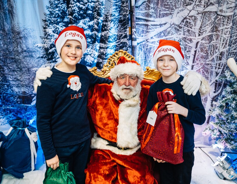 Two Children standing either side of Santa Claus in Santa's Grotto. They are each wearing Christmas hats.