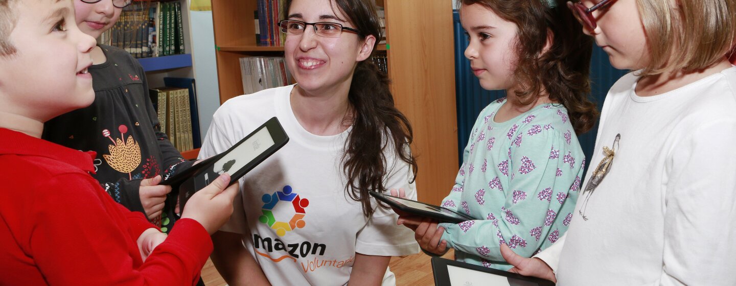 Una asociada de un centro logístico observa sonriendo la cara de sorpresa de cinco alumnos de una escuela de primaria beneficiaria de una Kindle Donation. Ella está arrodillada y los cuatro pequeños tienen un Kindle entre las manos.
