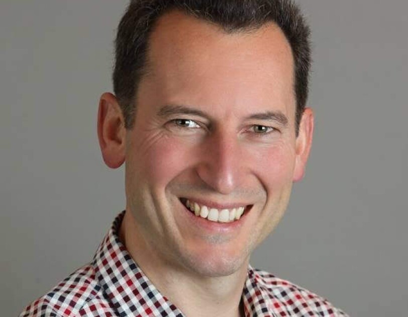 Alex Smola, machine learning director for Amazon Web Services’ Deep Engine group wears a blue striped hooded sweatshirt over a black tee shirt, and a cross-body bag. He stands outside near a rock formation.