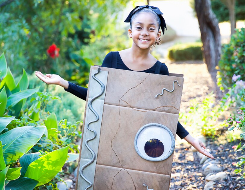 A young girl wears a costume made of an Amazon box, decorated to look like a spell book with a giant eye and silver snakes. The box is attached with shoulder straps and covers from her shoulders to her knees. The