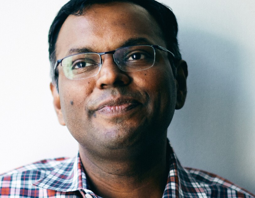 A close-cropped image of a man wearing a plaid button up over a white tee, and glasses sits in front of a white wall.