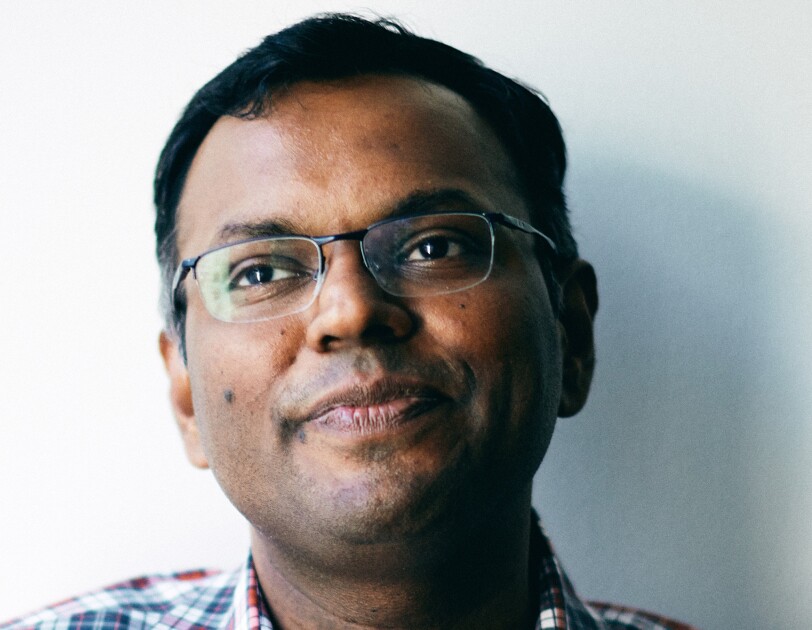 A close-cropped image of a man wearing a plaid button up over a white tee, and glasses sits in front of a white wall.
