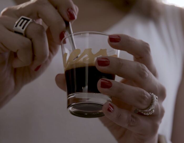 Primo piano di due mani femminili che reggono una tazzina di caffè in vetro. Sulla tazzina si intravede il logo 'Yespresso'. La donna, di cui non si vede il volto, indossa una canotta bianca, ha due anelli su entrambi gli anulari e lo smalto rosso. La sua mano sinistra regge la tazzina, mentre con la destra gira il caffè con un cucchiaino.