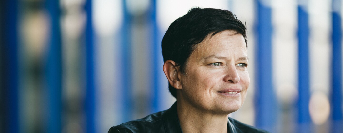 A dark-haired woman, Amazon's disaster relief head Bettina Stix, is dressed in a black leather jacket looks to the right and smiling. There is a blue background.
