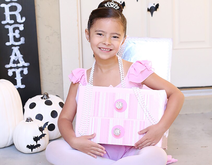 A little girl in a costume made from an Amazon box. The box is crafted to look like a jewelry box. The girl is wearing ballet slippers, tights, a leotard, pearl necklaces and a tiara. The costume is a "jewelry box ballerina"