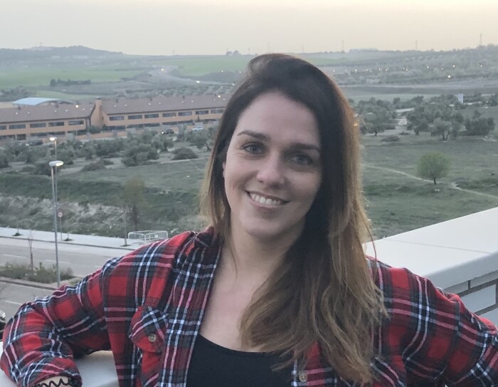 Noemi Cameselle. Con los ojos azules y el pelo liso y por los hombros de color castaño con mechas rubias, Noemí está mirando a cámara con una leve sonrisa. Va vestda con una camiseta negra y por encima un jersey camisero de cuadros negros, rojos y blancos. Está situada en una terraza y de fondo se ve una caretera de dos carriles por cada senido y viviendas al fondo.