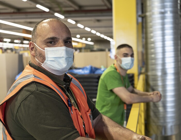Santiago And´res y Santi Andrés en el centro logístico de Amazon en San Ferando de Henares (Madrid). Desenfocado, de fondo, las estanterías amarillas de un centrlo logístico. En primer plano el padre, Santiago Andrés con pelo muy corto negro y blanco y un poco calvo. Lleva un polo de manga corta de color verde, chaleco naranja y mascarilla. Su hujo en segundo plano con camiseta verde más destacada y el badge con una cinta naranja y mascarilla. Los dos están apoyados a la barandilla amarilla.