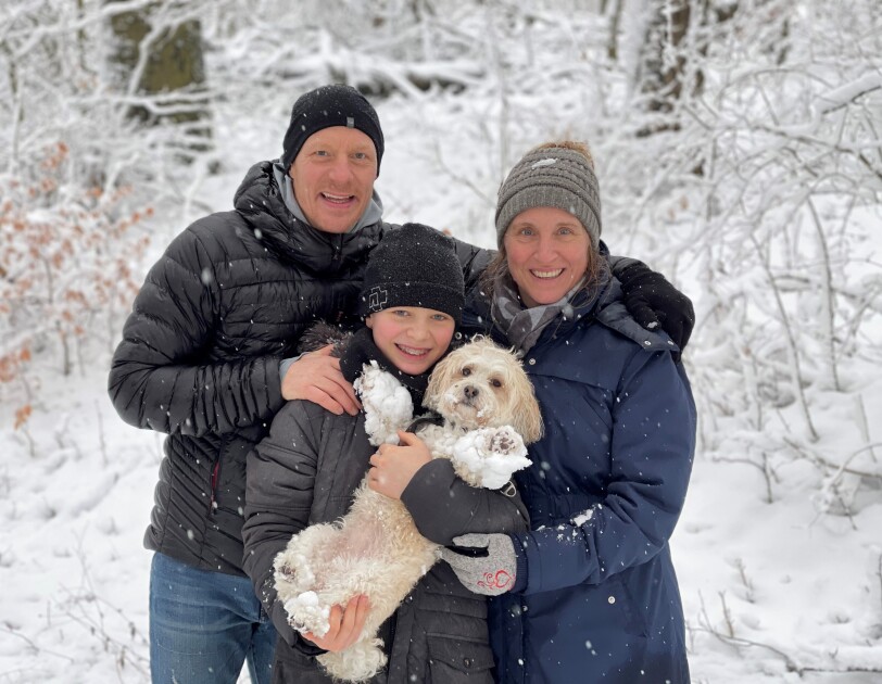 Ein Mann, eine Frau und ein Kind mit Hund in den Armen stehen im Schnee und lachen in die Kamera.