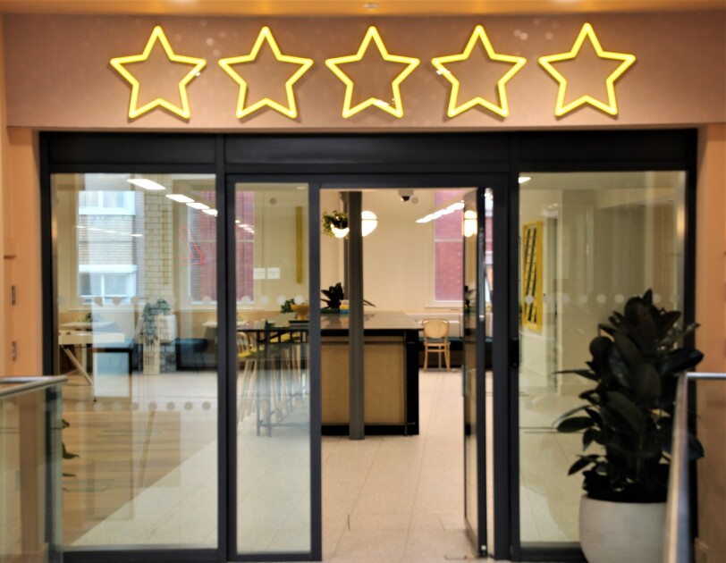 A view of the break area in the Amazon Manchester offices, this is viewed from outside of the open doors. Above the doors are neon lights depicting five stars.