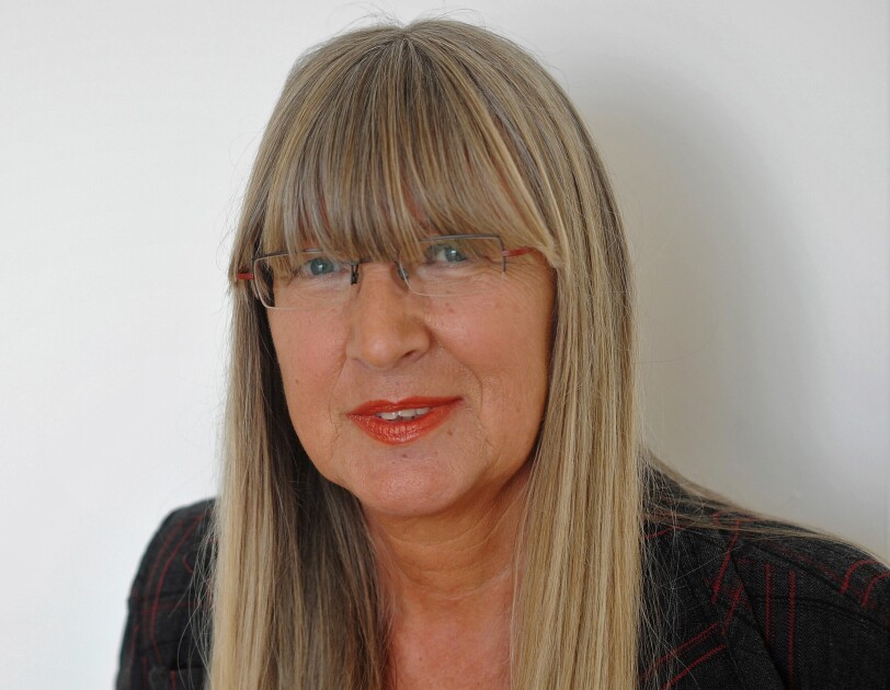 Eine Frau mit blonden langen Harren und Brille lächelt in die Kamera. Sie trägt roten LIppenstift.