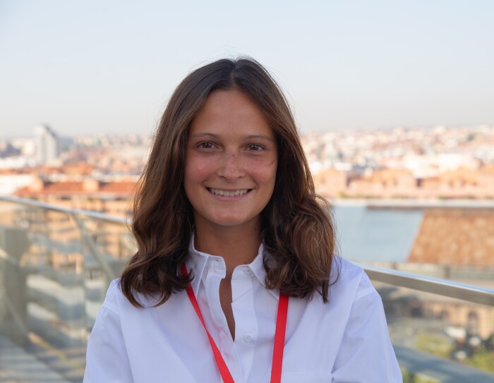María Robert, Senior Program Manager de Experiencia del Cliente. De fondo el paisaje de Madrid. Está en el último piso de las oficinas corporativas. Hay una barandilla de color metal. María tiene el pelo castaño un poco más allá del hombro. Lo lleva suelto. Está morena, tiene pecas y lleva una camisa blanca.