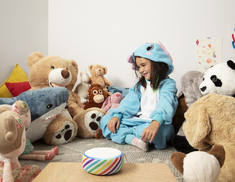 A child sits in a playroom, surrounded by stuffed animals. In front of her is an Echo Dot device is a rainbow stripe, on a tabletop.
