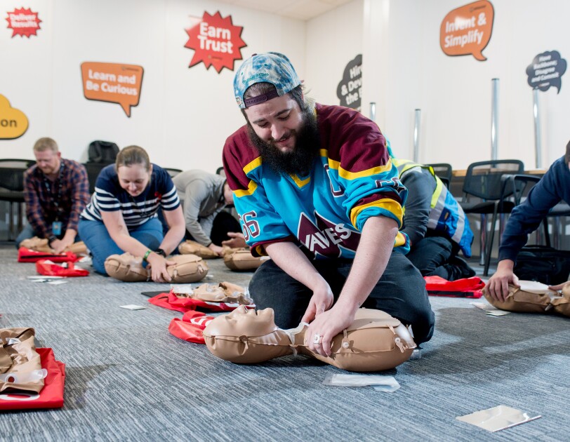 Amazon Operations Supervisor Lloyd Curley kneeling on the floor and practicing CPR on a dummy, along with five of his colleagues doing the same in the background.