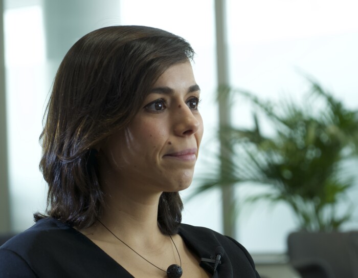 Natalia Gutiérrez, Líder Global de Impacto Sostenible y Seguridad de Cabify. De fondo unas butacas de color gris con plantas verdes y una lámpara de pie de color blanco. Unos ventanales grises grandes. La protagonista en primer plano con una camisa cruzada negra de cuello pico, manga larga y botones de color blanco. Con el pelo castaño, liso hasta el hombro y sonriendo mirando a la derecha de la foto. Lleva un collar de alambre con una bola.