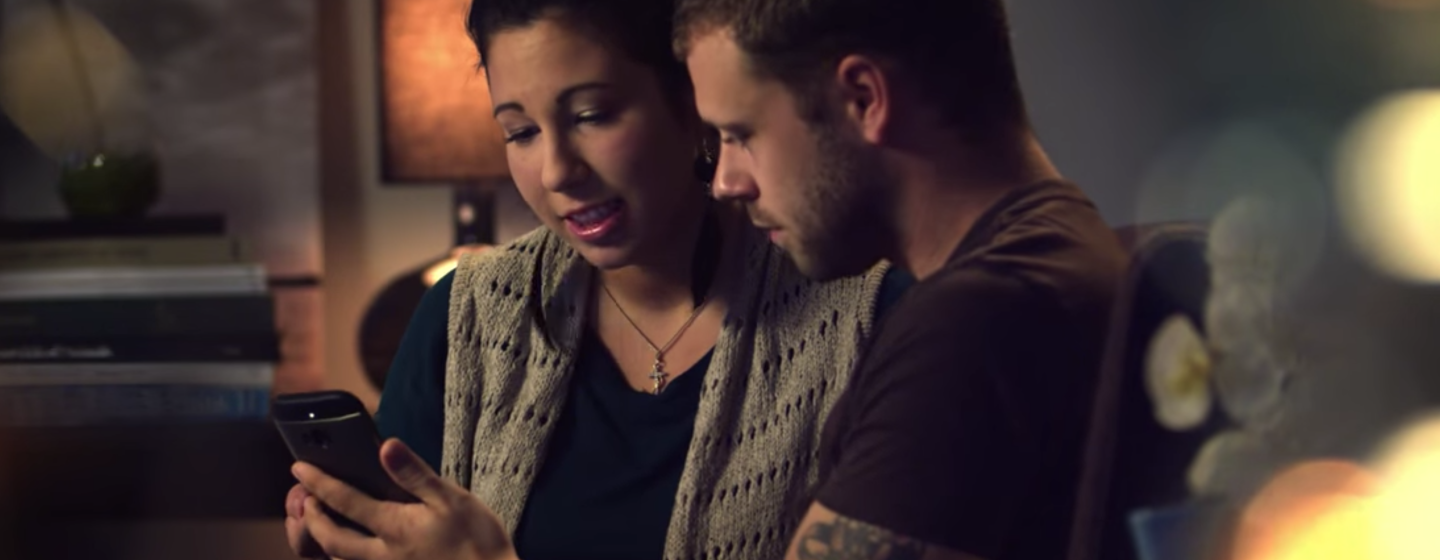Un uomo e una donna guardano lo schermo di uno smartphone nella stanza di un'abitazione. L'uomo regge lo smartphone con il braccio sinistro. Alla sua destra, la donna gli indica lo schermo con la mano destra.