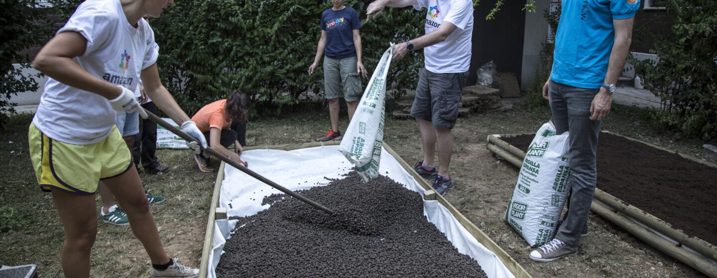 A scuola di verde: sei volontari Amazon all'opera nel giardino della scuola Media Leonardo da Vinci di Affori, nella periferia di Milano. Una donna in primo piano regge una pala con cui stende della terra, versata da un sacco da un uomo, in piedi di fronte alla donna. Gli altri 4 volontari osservano la scena.