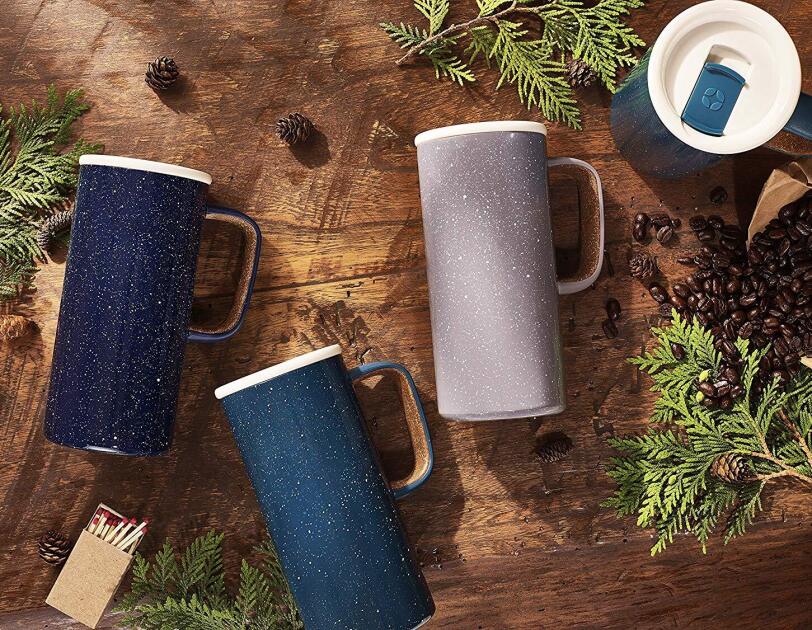 Three tall travel mugs lie on their side, on a rustic wood backdrop. They're surrounded by pine branches, pine cones, and coffee beans. A fourth mug stands in the upper right corner, sealed with a lid.