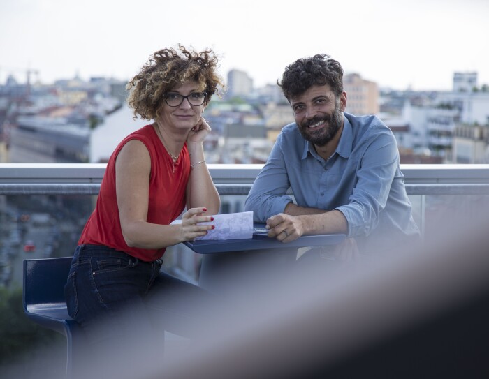 Due persone siedono allo stesso tavolo, sulla terrazza del palazzo Amazon di Milano. A destra, Alessandro Saccon, Consumer PR Manager. Di fronte a lui siede Alessandra Meucci, Executive Assistant. Sullo sfondo lo skyline di Milano. La foto è stata scattata in occasione del Pride di Milano di giugno 2018.
