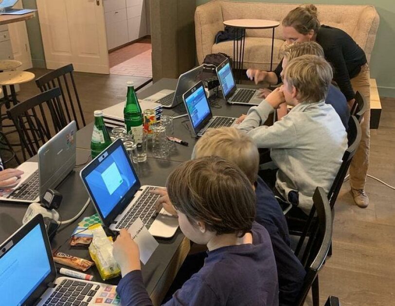 digital.engagiert_oderdojo_Kinder sitzen an einem langen Tisch, vor ihnen stehen Laptops. Eine Frau erklärt ihnen etwas.
