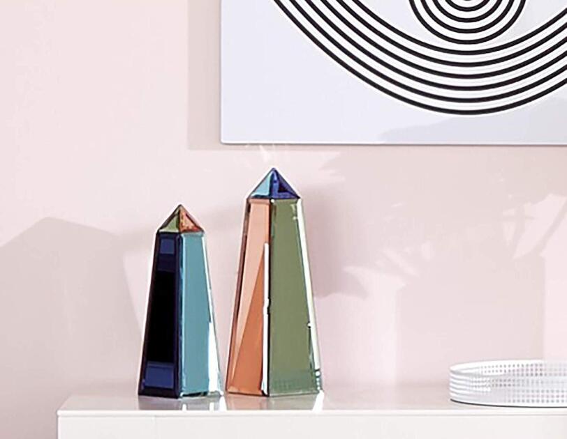 Two multi-color obelisks on a lacquered table. The wall behind the items is soft pink, with a black and white art print depicting an eye.