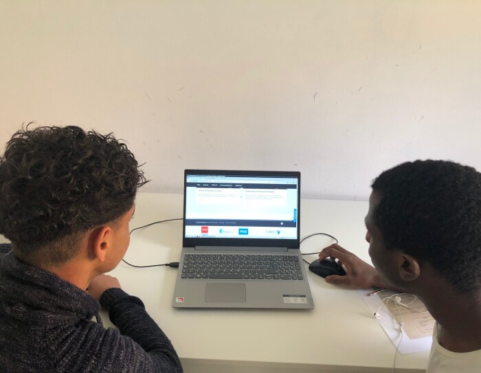 La donación de Amazon a la Fundación Diagrama ha sido de 39 ordenadores portátiles y 72 tabletas. En la foto aparecen dos chicos negros en primer plano con el peli también negro. Unos lleva una camisa negra y el otro una camiseta blanca. Entre ellos hay un ordenador portátil encendido y el chico de la derecha sujeta el ratón con la mano. La mesa es de color blanco y la pared también.
